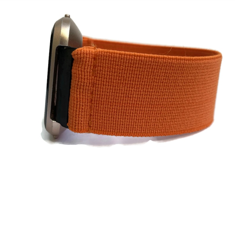 K&ouml;nnte beinhalten: Eine silberne Smartwatch mit einem orangefarbenen Stoffarmband. Das Armband ist aus gewebtem Material und hat eine glatte Textur.