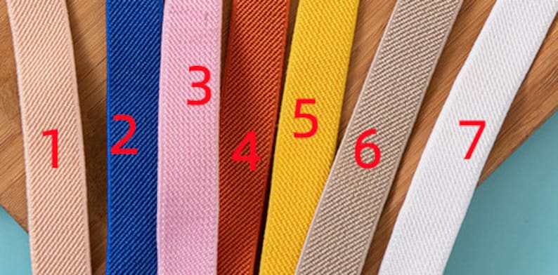 May include: Seven different types of elastic ribbon in various colours, including beige, blue, pink, orange, yellow, and white. The ribbon is arranged horizontally on a wooden surface.
