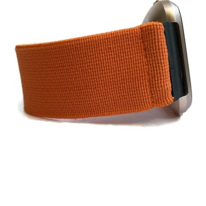 K&ouml;nnte beinhalten: Orangefarbenes Armband aus gewebtem Stoff. Das Armband ist an einem silbernen Zifferblatt befestigt.