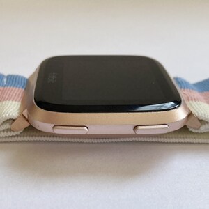May include: A rose gold smartwatch with a black screen and two buttons on the side. The watch is on a white and blue striped elastic band.