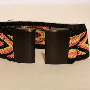 Op de afbeelding: Een zwart elastiek met een roze en geel geometrisch patroon. Het elastiek heeft twee zwarte plastic gespen.