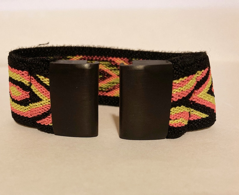 Op de afbeelding: Een zwart elastiek met een roze en geel geometrisch patroon. Het elastiek heeft twee zwarte plastic gespen.
