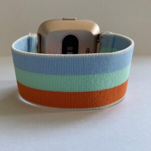 May include: A gold-colored smartwatch with a blue, green, and orange striped elastic band.