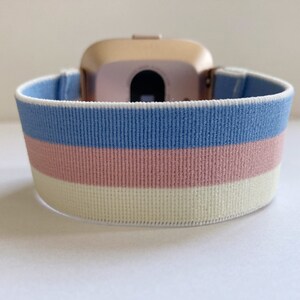 May include: A light blue, pink, and white striped elastic watch band. The band is made of a soft, stretchy material and is designed to fit comfortably on the wrist.