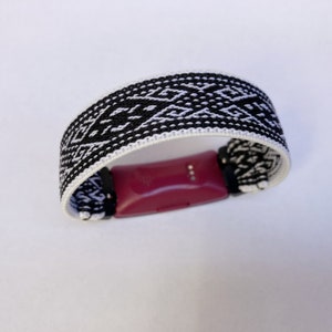 Peut inclure: Un bracelet tiss&eacute; noir et blanc avec un motif g&eacute;om&eacute;trique, con&ccedil;u pour maintenir un tracker de fitness bordeaux. Le bracelet mesure environ 2,5 cm de large et pr&eacute;sente un motif de losanges r&eacute;p&eacute;t&eacute;s. Le tracker est maintenu par des connecteurs noirs.