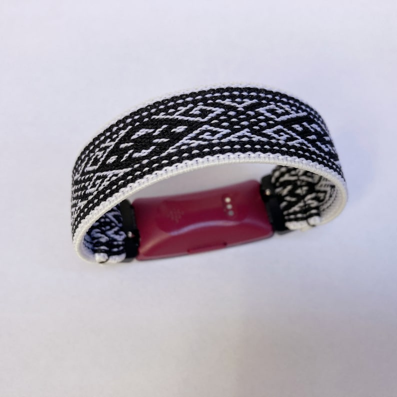 Peut inclure: Un bracelet tiss&eacute; noir et blanc avec un motif g&eacute;om&eacute;trique, con&ccedil;u pour maintenir un tracker de fitness bordeaux. Le bracelet mesure environ 2,5 cm de large et pr&eacute;sente un motif de losanges r&eacute;p&eacute;t&eacute;s. Le tracker est maintenu par des connecteurs noirs.