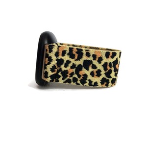 May include: A black and tan leopard print watch band. The band is made of a stretchy material and has a black buckle.