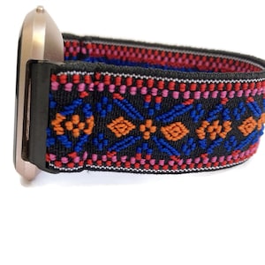 Op de afbeelding: Een zwart horlogebandje met een kleurrijk geometrisch patroon in rood, oranje, blauw en roze. Het bandje is gemaakt van rekbaar materiaal en heeft een goudkleurige metalen gesp.