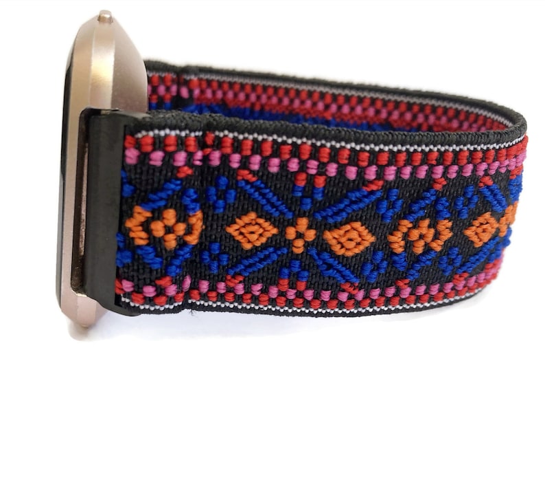 Op de afbeelding: Een zwart horlogebandje met een kleurrijk geometrisch patroon in rood, oranje, blauw en roze. Het bandje is gemaakt van rekbaar materiaal en heeft een goudkleurige metalen gesp.
