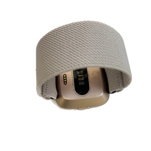 May include: A gold-colored fitness tracker with a gray band. The back of the tracker shows the text "Aerobic Grade Aluminum Case", "Optical Heart Rate", "Designed at Fitbit", and "CE".