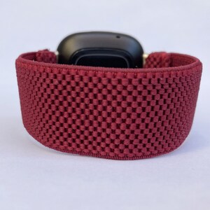 May include: A close-up of a maroon sport band for a smartwatch. The band is made of a soft, breathable material with a textured pattern.