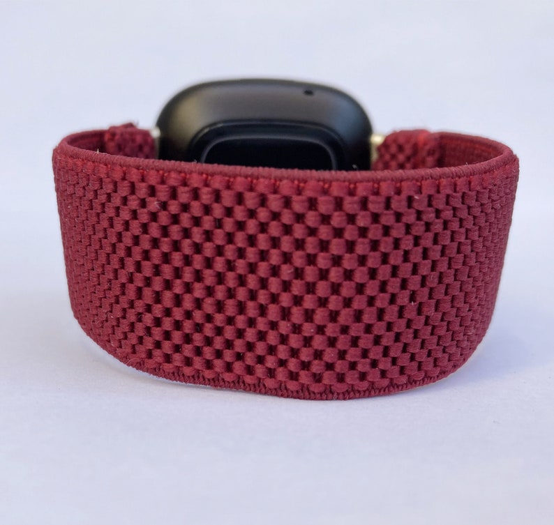 May include: A close-up of a maroon sport band for a smartwatch. The band is made of a soft, breathable material with a textured pattern.