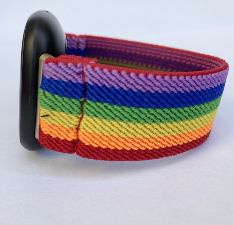 May include: A rainbow-striped watch band with a black watch face. The band features horizontal stripes in red, orange, yellow, green, blue, and purple. The band is made of a woven material.