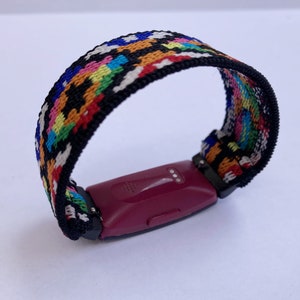 Op de afbeelding: Een kleurrijk geweven bandje met patroon voor een fitness tracker. Het bandje is zwart met een veelkleurig geometrisch patroon. Het bandje is bevestigd aan een bordeauxrode fitness tracker.