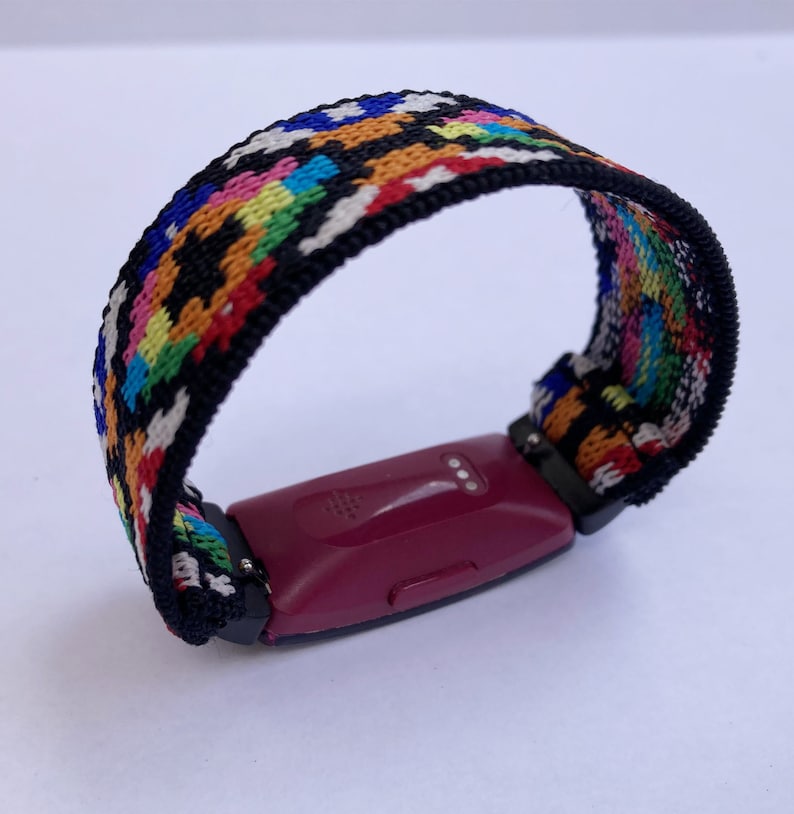 Op de afbeelding: Een kleurrijk geweven bandje met patroon voor een fitness tracker. Het bandje is zwart met een veelkleurig geometrisch patroon. Het bandje is bevestigd aan een bordeauxrode fitness tracker.