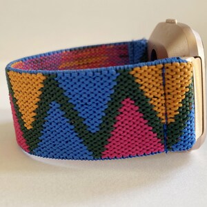 May include: A gold-colored smartwatch with a colorful geometric patterned band. The band is made of a woven fabric with blue, pink, yellow, and green triangles.