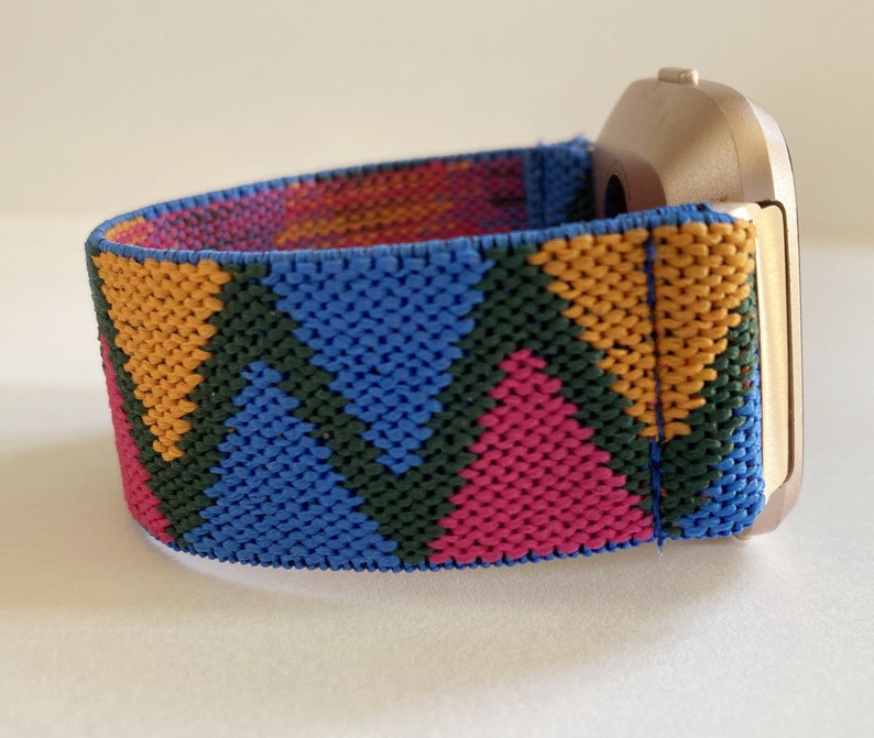 May include: A gold-colored smartwatch with a colorful geometric patterned band. The band is made of a woven fabric with blue, pink, yellow, and green triangles.