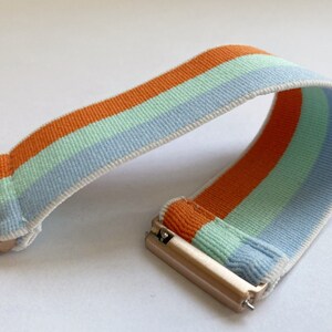 May include: A striped watch band with orange, light blue, and light green stripes. The band is made of a soft, stretchy material and has a gold metal clasp.