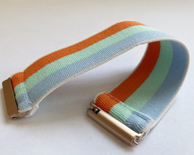 May include: A striped watch band with orange, light blue, and light green stripes. The band is made of a soft, stretchy material and has a gold metal clasp.