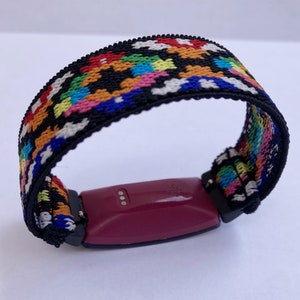 May include: A fitness tracker with a colourful woven band. The band is black with red, orange, yellow, green, blue and white stripes. The tracker is a dark purple colour and has three small silver dots on the side.