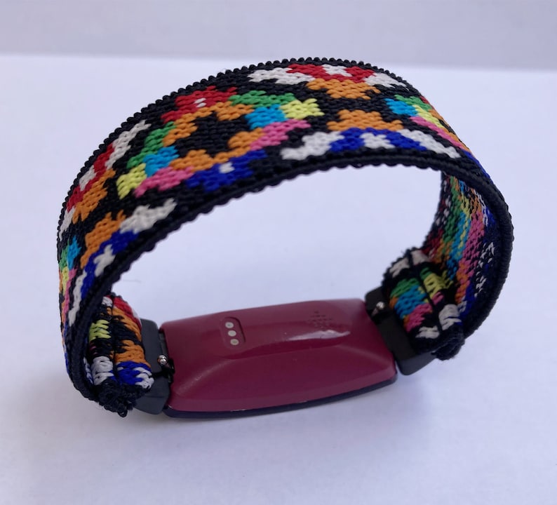 May include: A fitness tracker with a colourful woven band. The band is black with red, orange, yellow, green, blue and white stripes. The tracker is a dark purple colour and has three small silver dots on the side.