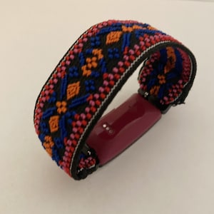 Op de afbeelding: Een roze fitness tracker met een kleurrijk geweven bandje. Het bandje heeft een geometrisch patroon in tinten blauw, oranje, roze en zwart.