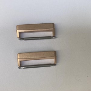 May include: Two gold-colored watch band spring bars. The spring bars are made of metal and have a silver finish.