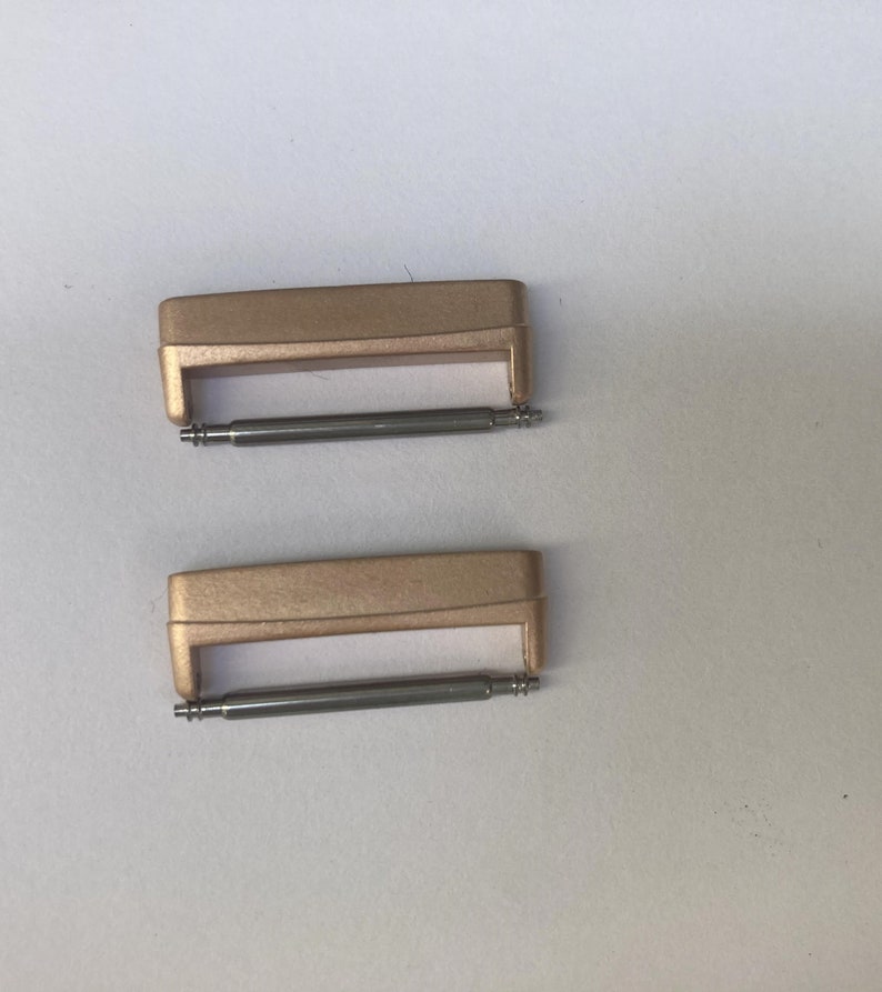 May include: Two gold-colored watch band spring bars. The spring bars are made of metal and have a silver finish.
