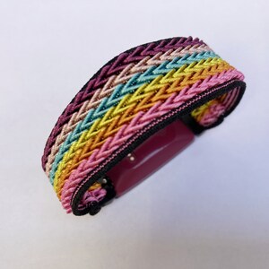 Op de afbeelding: Een kleurrijke geweven armband met een regenboogpatroon. De armband heeft in elkaar verweven draden in tinten roze, geel, turquoise en paars, met een zwarte rand. De sluiting heeft een rechthoekige vorm in een dieproze kleur.