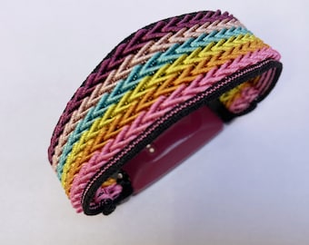 Puede incluir: Una colorida pulsera tejida con un patrón de arcoíris. La pulsera presenta hilos entrelazados en tonos de rosa, amarillo, turquesa y morado, con un borde negro. El cierre tiene forma rectangular y es de color rosa intenso.
