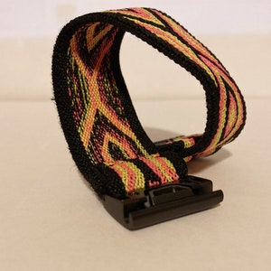 Op de afbeelding: Een zwart horlogebandje met een roze en geel gestreept patroon. Het bandje heeft een zwarte metalen gesp.