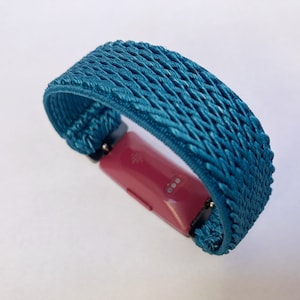 Op de afbeelding: Een geweven band in turquoise blauw voor een fitness tracker. De band is gemaakt van een zacht, flexibel materiaal en heeft een textuur. De band is ontworpen om comfortabel te dragen en is compatibel met de meeste fitness trackers.