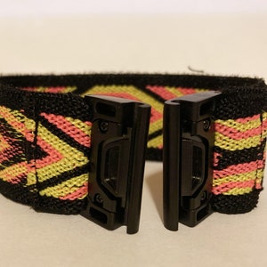 Op de afbeelding: Zwart elastisch horlogeband met een geometrisch patroon in roze en geel. Het band heeft twee zwarte metalen gespen.