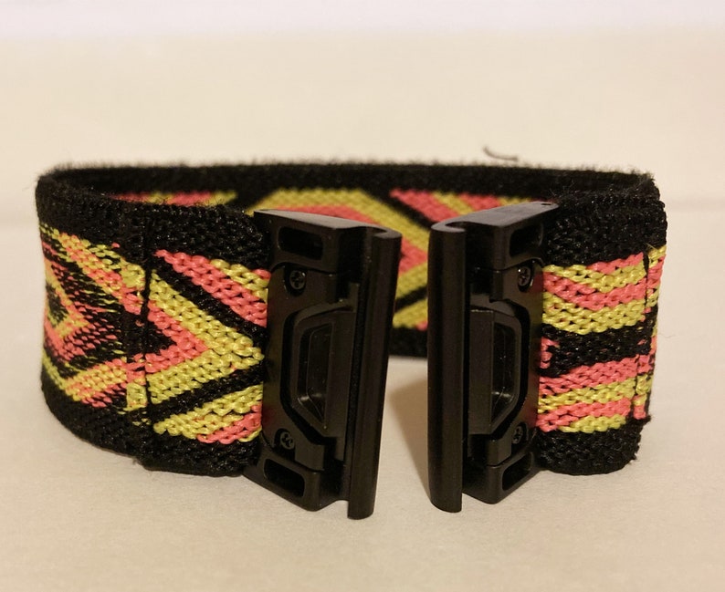 Op de afbeelding: Zwart elastisch horlogeband met een geometrisch patroon in roze en geel. Het band heeft twee zwarte metalen gespen.