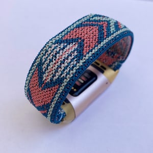 May include: A pink, blue and white patterned woven band for a fitness tracker. The band is attached to a gold-coloured fitness tracker.