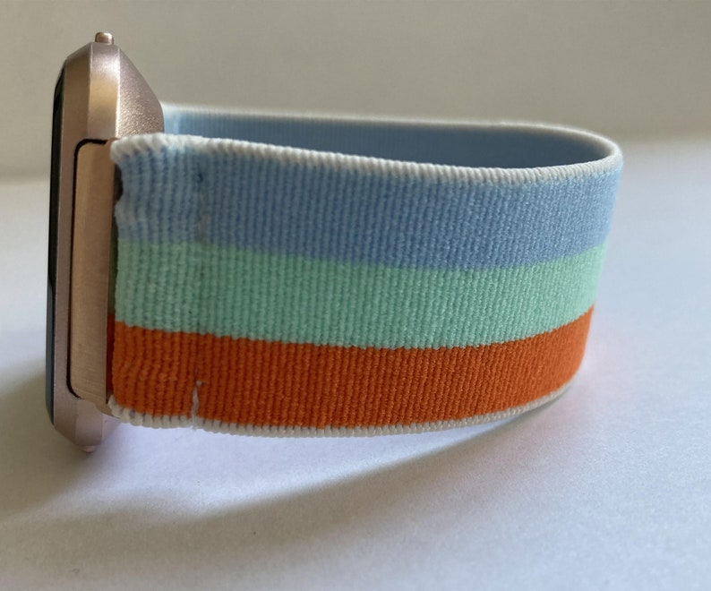 May include: A rose gold smartwatch with a striped blue, green, and orange elastic band.