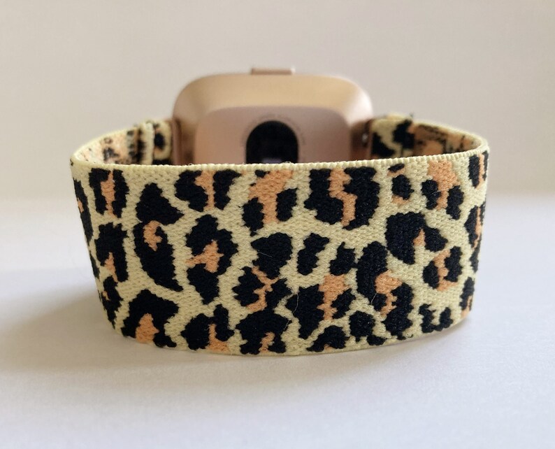 May include: A light beige elastic watch band with a leopard print pattern in black and orange. The band is designed for a smartwatch.