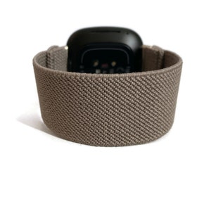 K&ouml;nnte beinhalten: Ein braunes elastisches Armband mit einem schwarzen Zifferblatt. Das Armband besteht aus einem weichen, dehnbaren Material und hat eine strukturierte Oberfl&auml;che.