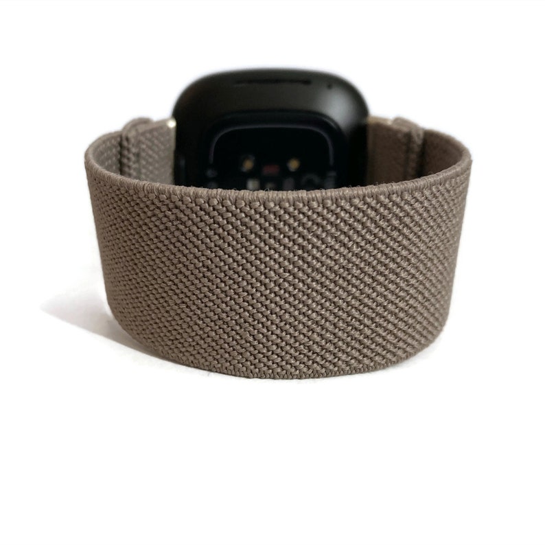 K&ouml;nnte beinhalten: Ein braunes elastisches Armband mit einem schwarzen Zifferblatt. Das Armband besteht aus einem weichen, dehnbaren Material und hat eine strukturierte Oberfl&auml;che.