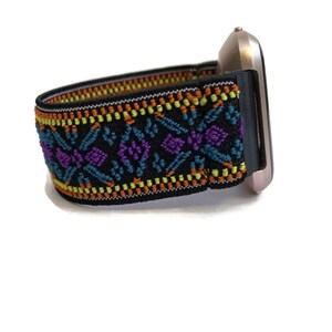K&ouml;nnte beinhalten: Ein schwarzes elastisches Armband mit einem bunten geometrischen Muster in Orange-, Gelb-, T&uuml;rkis-, Lila- und Wei&szlig;t&ouml;nen. Das Armband ist an einer silbernen Smartwatch befestigt.