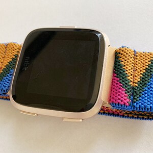 以下が含まれることがあります： ローズゴールドの Fitbit スマートウォッチとカラフルな織りバンド。バンドは青、緑、ピンク、黄色の幾何学模様です。