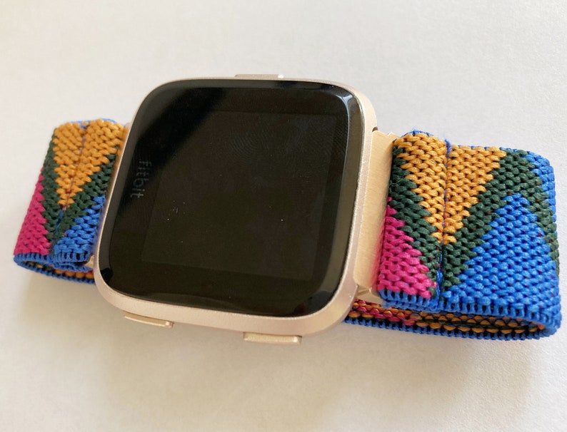 以下が含まれることがあります： ローズゴールドの Fitbit スマートウォッチとカラフルな織りバンド。バンドは青、緑、ピンク、黄色の幾何学模様です。