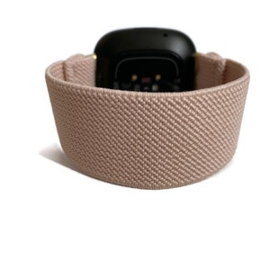 May include: A light pink elastic watch strap with a black watch face. The strap is made of a soft, textured material.