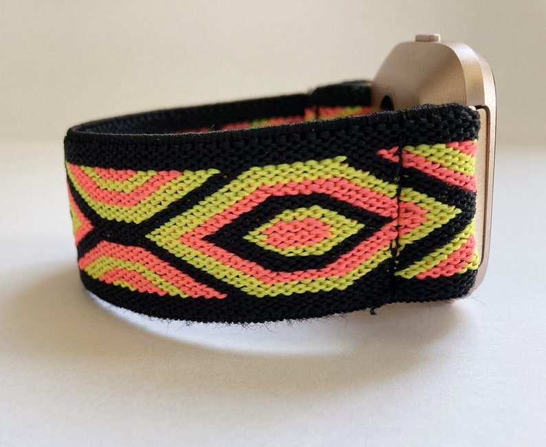 May include: A black elastic watch band with a geometric pattern in pink and green. The band is attached to a silver watch face.
