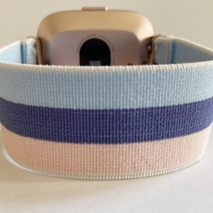 May include: A light blue, dark blue and pink striped elastic watch strap. The strap is made from a soft, stretchy material and is designed to fit most wrist sizes.