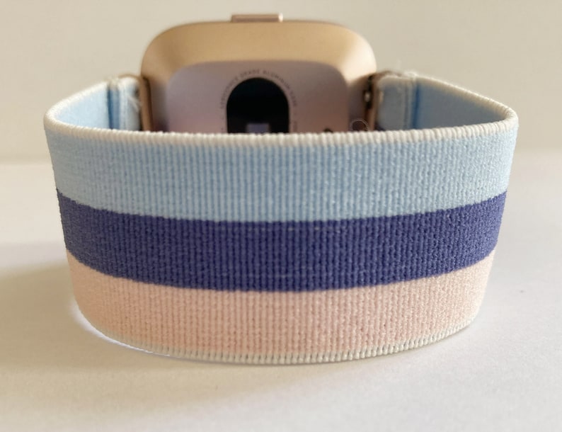 May include: A light blue, dark blue and pink striped elastic watch strap. The strap is made from a soft, stretchy material and is designed to fit most wrist sizes.