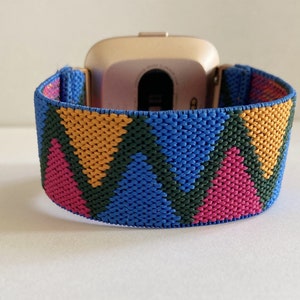 May include: A gold smartwatch with a colorful geometric patterned band. The band is made of woven fabric and features pink, blue, green, and yellow colors.