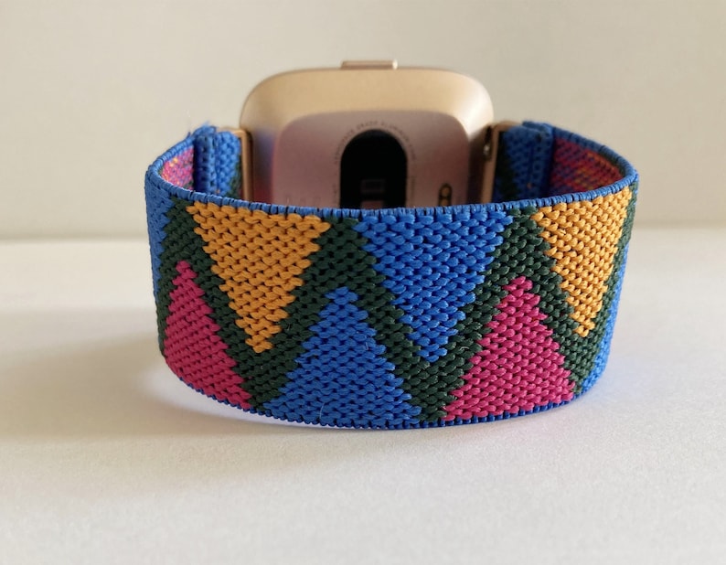 May include: A gold smartwatch with a colorful geometric patterned band. The band is made of woven fabric and features pink, blue, green, and yellow colors.