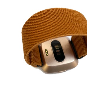 May include: A gold-colored fitness tracker with a brown fabric band. The tracker has a small screen and a few buttons on the side.