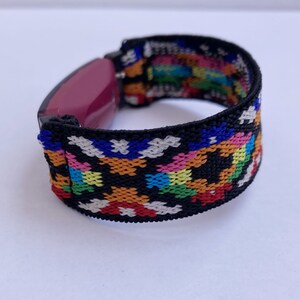 May include: A black and colourful patterned elastic band for a fitness tracker. The band is made of woven fabric with a geometric design in red, orange, yellow, green, blue, pink, and white.